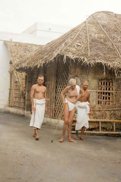 photo of Sri Bhagavan walking with two attendants in front of a thatched shed