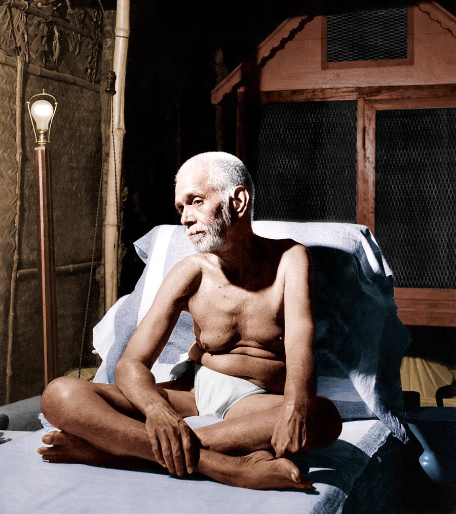 colour photo of Sri Bhagavan seated cross-legged on a bed with a lamp and wooden window frame behind