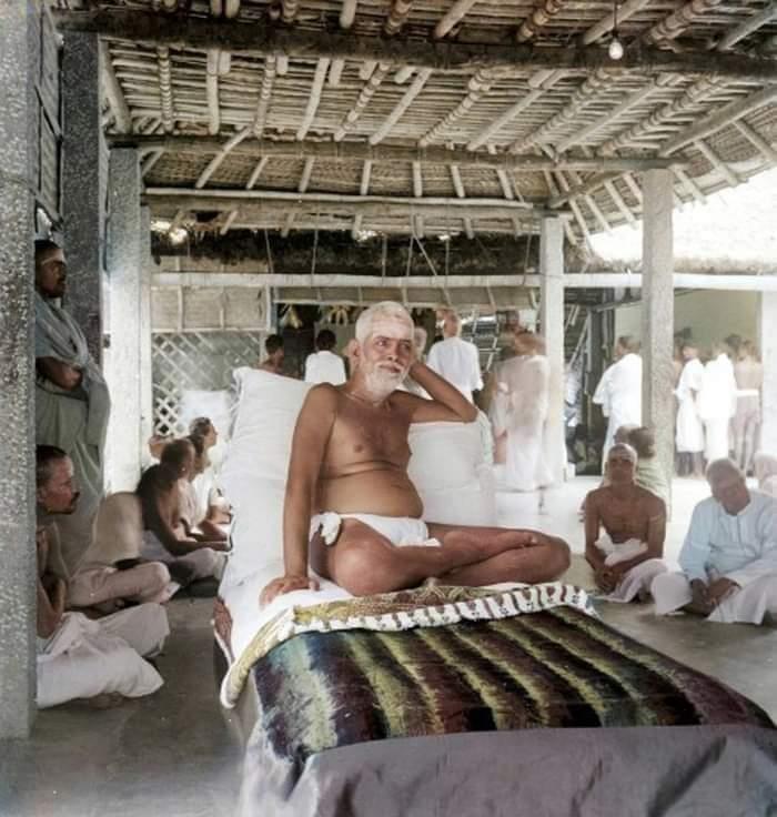colour photo of Sri Bhagavan seated on a couch under a thatched roof, devotees seated around