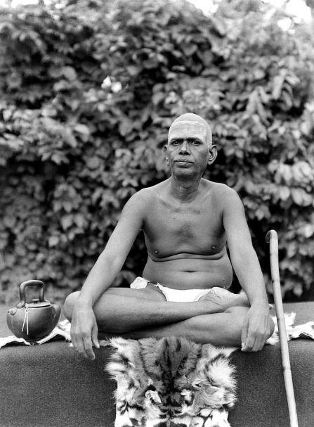 photo of Sri Bhagavan seated in padmasana on a tiger skin in front of a leafy tree, water pot at left, walking stick at right