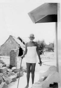 photo of Sri Bhagavan standing with a stick beside a thatched shed