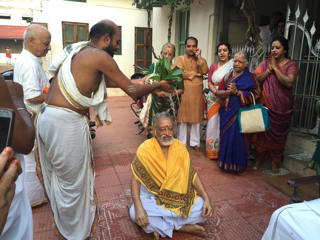 priest pouring water on V. Ganesan seated on floor in yellow shawl, onlookers behind
