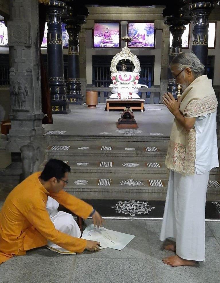 Dr. Anand seated on floor signing document before shrine, V. Ganesan standing with folded hands