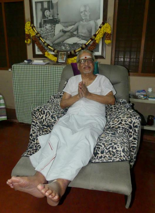 elderly man in white seated in armchair with folded hands, framed photo of Sri Bhagavan behind
