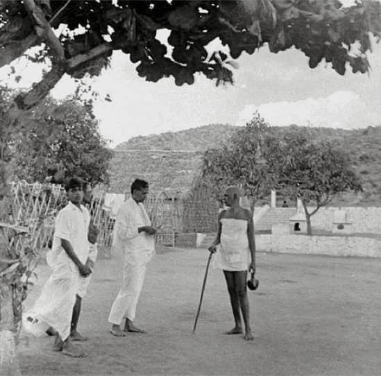 photograph of Sri Bhagavan standing with staff and water pot, talking with two men in white in an open yard beneath a tree, hill behind