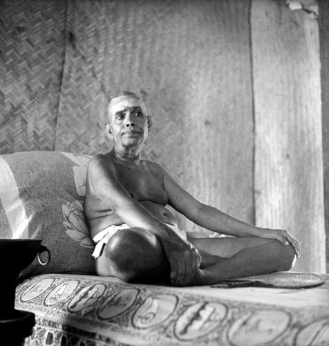 photograph of Sri Bhagavan seated on the couch, one knee raised, looking to his left; no caption