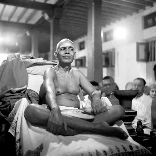 photo of Sri Bhagavan seated cross-legged on a couch, devotees behind; no caption