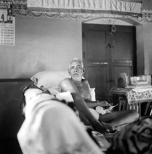 photo of Sri Bhagavan reclining on a couch, bookshelf and door behind; no caption